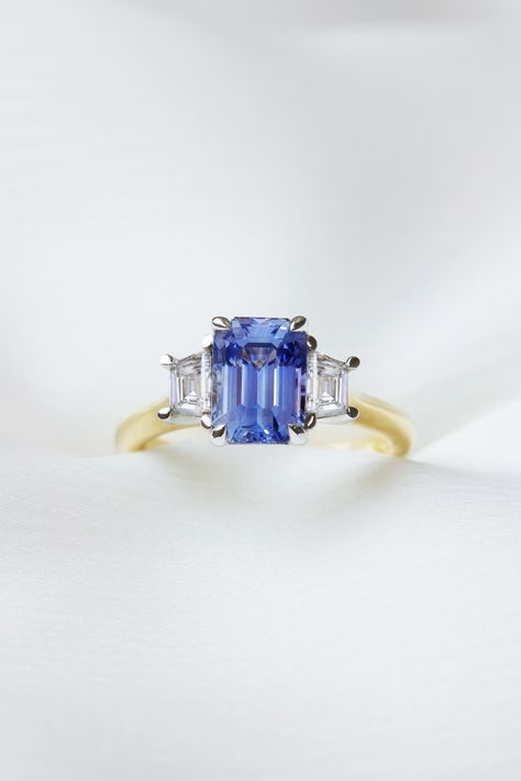 Featuring a vibrant, 2.12ct emerald cut sapphire accented by two trapezoid-cut diamond side stones, this bespoke 18ct yellow gold trilogy ring flawlessly fuses contemporary colour with old world glamour. Luxury Three Stone Sapphire Ring With Baguette Cut, Timeless Sapphire Baguette Cut Ring, Emerald Cut Three Stone Sapphire Ring, Sapphire Trilogy Engagement Ring, Whie And Blue Trrilogy Sapphire Engagement Ring, Sapphire And Diamond Trilogy Ring, Sapphire Trilogy Ring, Sapphire Ring Designs, Old World Glamour