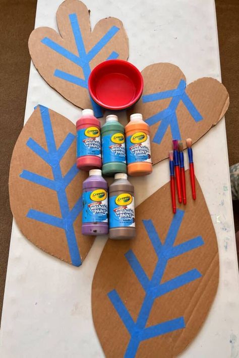 A setup of materials for the craft on a table, including cardboard leaves with painter’s tape in leaf vein patterns, bright-colored paint bottles, paintbrushes, and a red bowl, ready for a fall-themed preschool activity. Cardboard Leaves, Preschool Fall Art, Diy Thanksgiving Centerpiece, Leaf Crafts Kids, Thanksgiving Centerpieces Diy, Paint Bottles, Cardboard Creations, Leaf Craft, Preschool Fall