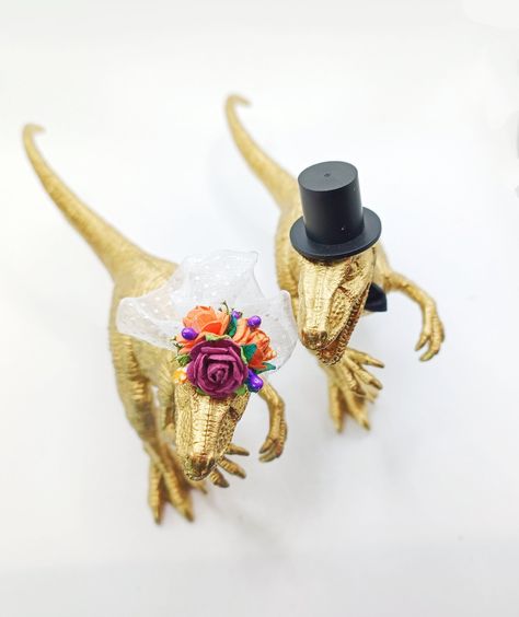 "Our eye catching gold dinosaur wedding cake toppers add a completely individual touch to your special day.  The dino bride comes with a veil and flower crown, while the groom has a top hat and bow tie!  As standard, we provide a Velociraptor bride and T Rex groom, but can do any combination you would like. Just let us know in the personalisation section. We can also source other Dino types if you would prefer!  Our aim is to work with you to create completely bespoke dinos to match your theme, so be as unique as you like! We can create different colour dinos and give them a variety of eclectic accessories such as crowns, pearls, tutus and a range of fun items such as bottles/glasses of wine, mini cake slices and cans of beer. For these options or any ideas of your own, just send us a mess Dinosaur Wedding Cake Topper, Fun Cake Toppers Wedding, Gold Dinosaur Cake, Gay Wedding Ideas Decor, Dinosaur Wedding Theme, Dino Wedding, Dinosaur Wedding Cake, Wedding Cake Topper Gold, Wedding Decor Gold