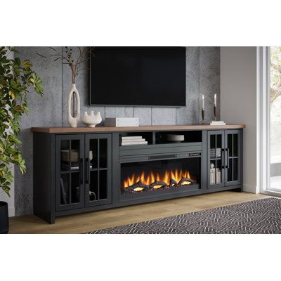 This TV stand comes with an electric fireplace insert that adds a cozy ambiance to your living room. A remote is included to control the heat output, the LED flickering flame effect, and the timer. Crafted from solid wood with a contrasting two-tone finish, this stand lends a modern farmhouse vibe thanks to its clean lines and the windowpane paneling on both cabinet drawers. Glass fronts let you look inside at your DVD or game collection, while two cubbies over the insert are ideal for consoles Tv Console With Fireplace And Storage, Rustic Entertainment Center With Fireplace, 98 Inch Tv Living Room, Living Room With Electric Fireplace Tv Stand, Electric Fireplace Tv Stands, Electric Fireplace Furniture, Fake Fireplace Tv Stand, Fireplace Console Decor, Gothic Tv Stand