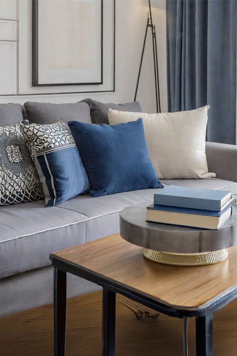 Refined Comfort • Chic living room corner with soft, textured drapery • Elegant grey sofa adorned with an array of decorative cushions • Mixture of bold blue and intricate patterned pillows adds visual interest • Contemporary round metal coffee table with a golden base • A stack of hardcover books atop the table suggests a quiet reading nook • Nested wooden side table introduces a functional layer to the design • Neutral-toned artwork and floor lamp complement the room's modern aesthetic. Gray And Blue Aesthetic, Blue Aesthetic Home Decor, Midmod Boho, Blue Aesthetic Home, Modern Grey Living Room, Living Room Gray, Round Metal Coffee Table, Patterned Pillows, Comfort Chic