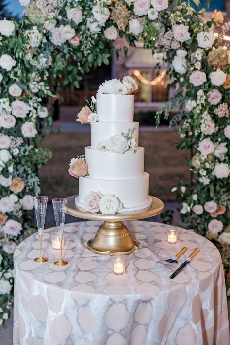 A beautiful white wedding cake adorned with delicate floral accents on a gold stand, surrounded by soft candlelight. The elegant cake table is set against a lush floral arch, creating a romantic ambiance perfect for a sophisticated outdoor wedding celebration. Wedding Cake Table Ideas, Stradley Davidson, Gold Cake Stand, Stunning Wedding Venues, Food Wedding, Flower Arch, Table Flower, Wedding Cake Table, Themed Wedding Cakes