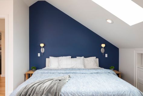 Attic main bedroom with sloped ceiling. It features a dark blue accent wall, sconces above the nightstands and an overhead skylight. Photo: Cindy Apple Photography Attic Accent Wall, Attic Bedroom Colors Sloped Ceiling, Tiny Attic Bedroom Sloped Ceiling, Bedroom With Sloped Ceiling Paint Colors, Dark Attic Bedroom, Sloped Ceiling Paint Ideas, Skylight Above Bed, Attic Paint Ideas Sloped Ceiling, Attic Kids Room Sloped Ceiling Beds