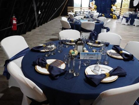 Set up a marvelous party ambiance and bedazzle your guests with your decorations. Drape navy blue curtains on a backdrop stand and bedeck them with white, gold, and blue balloons generating a cheerful appeal. Drape the tables with navy blue tablecloths and place lanterns in the middle for a gorgeous appeal. Moreover, spruce up the place settings using gold chargers, white plates, and navy blue napkins tied with rhinestone rings. Also, decorate the chairs with white covers and navy blue sashes. Navy Blue Table Settings, Gold And Blue Balloons, Navy Blue Table Cloth, White Wedding Table Setting, Navy Tablecloth, Navy Blue Napkins, Uc Merced, Party Ambiance, Event Decor Ideas