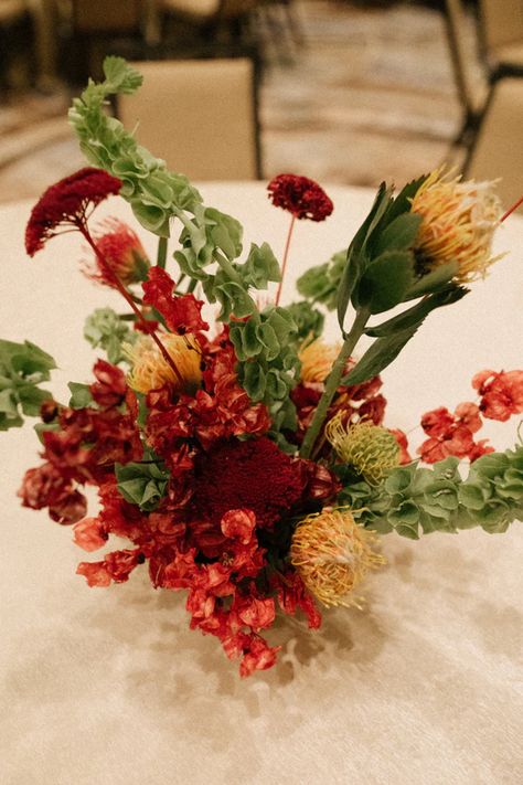 Unique floral centerpiece with lush green stems, deep red blooms and touches of electrifying yellow. Red And Green Centerpieces, Red And Green Flower Arrangements, Frank Lloyd Wright Style, Pincushion Protea, Coffee Table Arrangements, Bells Of Ireland, Taliesin West, Arizona Biltmore, Green Centerpieces