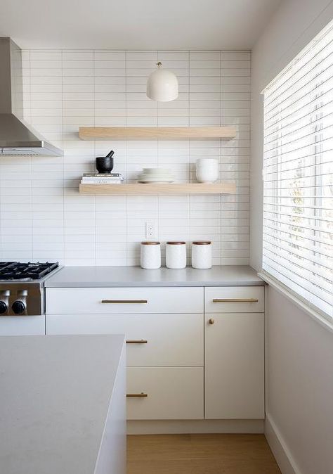 White flat front kitchen cabinets complemented with aged brass pulls are topped with a cement gray quartz countertop fixed against stacked glossy white backsplash tiles. Flat Front Kitchen Cabinets, Dapur Ikea, White Subway Tile Kitchen, Backsplash With White Cabinets, Oak Floating Shelves, White Tile Backsplash, Kabinet Dapur, Grey Countertops, Kitchen Backsplash Designs