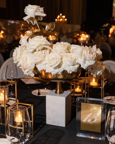 A sophisticated floral centerpiece designed by Ethereal Creators, featuring a lavish arrangement of white roses in a gold bowl, elegantly elevated on a white marble pedestal. The arrangement is accented with gold leaves, adding a touch of luxury. Surrounding the centerpiece are modern geometric gold candle holders, softly illuminating the table with warm candlelight. A gold and acrylic table number sign enhances the sleek, upscale aesthetic of the black-tabled decor Black White Gold Floral Centerpieces, Gold Floral Centerpieces, White Rose Centerpiece, Corporate Instagram, White Rose Centerpieces, Upscale Aesthetic, Table Number Sign, Rose Centerpiece, Marble Pedestal