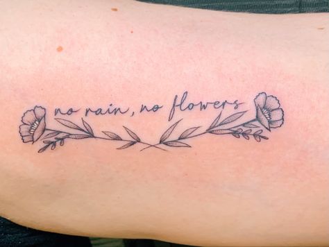 Script tattoo in font Artifact with two wispy flowers underneath. Tattoo reads “no rain, no flowers”. Flower Quote Tattoos For Women, Word Tattoo With Flowers Around It, No Rain No Flowers Wrist Tattoo, Flowers Surrounding Words Tattoo, Writing With Flower Tattoo, Lower Bicep Tattoo Women Words, It’s Ok Flower Tattoo, Pretty Script Tattoo, Flower Tattoo With Handwriting