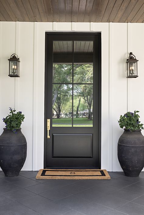 A white cottage home with board and batten trim is accented with a black front door finished with glass panels flanked by black lanterns and complemented with a monogrammed door mat. Black Front Door Glass Panel, Black Front Door With Glass Window, Front Door Privacy Glass Ideas, Front Door Glass Panel, Board And Batten Trim, Transitional Home Exterior, Front Door Transformation, White Rocking Chairs, Tiled Staircase