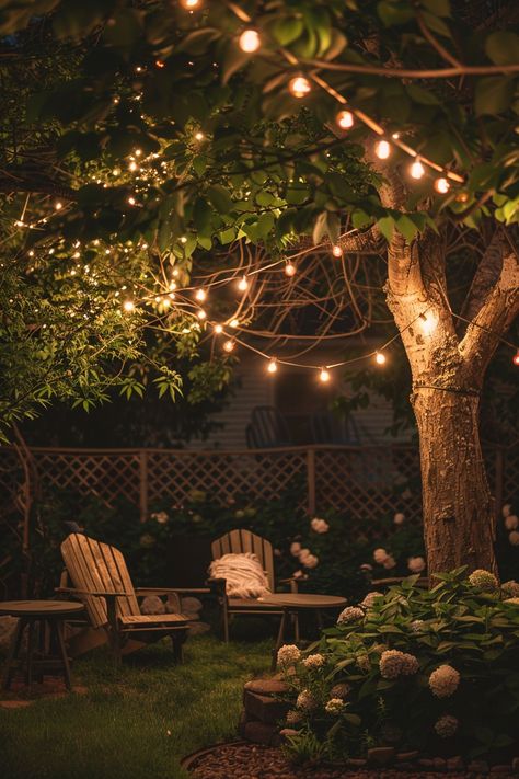 Cozy backyard patio with string lights illuminating wooden chairs surrounded by lush greenery and flowers, creating a perfect outdoor oasis. Bistro Lights Porch, Twinkle Lights In Trees, Fairy Lights Trees Backyards, Twinkly Lights Backyard, Lights In Trees Backyard, Ethereal Backyard, Yard Lighting Ideas, Backyard Lanterns, Cozy Backyard Ideas