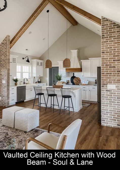 Brick columns frame this kitchen, which features white cabinets and countertops. Two woven pendant light fixtures hang from the vaulted ceiling above, which is accessorized with large wooden beams. Hardwood floors run the length of the room, while a white and wood hood vent is mounted on the back wall. 12ft Kitchen Ceiling, Small Kitchen With Vaulted Ceiling, High Ceiling Kitchen Ideas, Vaulted Ceiling Kitchen And Living Room, Low Ceiling Kitchen Ideas, Small Kitchen And Living Room Ideas, Small Kitchen And Living Room, Kitchen Vaulted Ceiling, Low Ceiling Kitchen