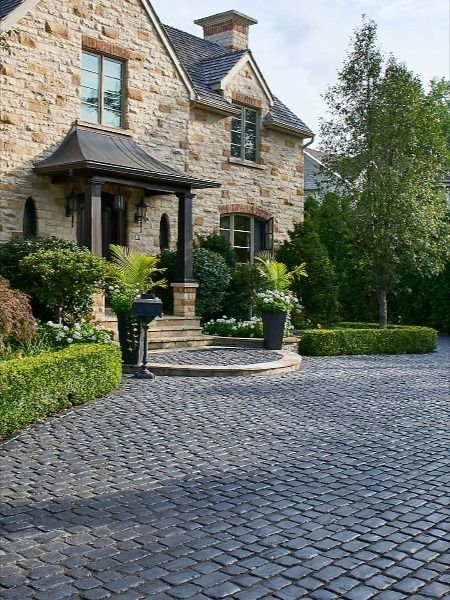 This European inspired home is brought to life with the charming cobblestone look of Courtstone. The dark tones of basalt are a beautiful, striking contrast to the beige bricks of the home. The deep green bushes, trees and grass add to the overall beauty of this landscape design. #FrontEntrance #Driveway #CurbAppeal #Courtstone Walk Path Ideas, Paved Patio Ideas, Cobbled Driveway, Pavers Walkway, Front Garden Ideas Driveway, Cobblestone Patio, Garden Ideas Driveway, Green Bushes, Driveway Entrance Landscaping