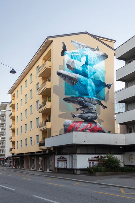 On the wall are painted several whales and sperm whales placed one on top of the other. In the upper part there is a blue parallelepiped that integrates with the composition of whales. In the lower part, on the whale that touches the ground, a red stripe is painted. Cube Architecture, Lugano Switzerland, Social Topics, Mural Paintings, Urban Intervention, Interactive Art, Human Condition, Mural Painting, Red Paint