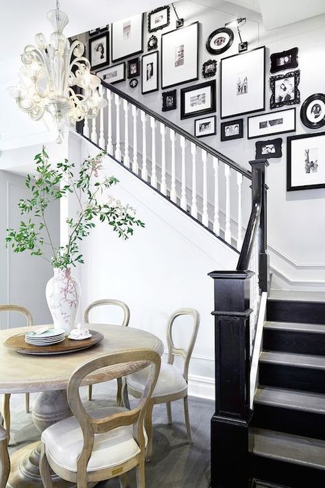 Foyer opens to black and white staircase accented with black and white photo wall alongside an eat-in kitchen boasting light wood round dining table surrounded by wood dining chairs. Black And White Hallway, Stairway Gallery, Stairway Gallery Wall, Family Photo Gallery Wall, Black And White Stairs, Stair Paneling, Picture Walls, White Hallway, Staircase Wall Decor