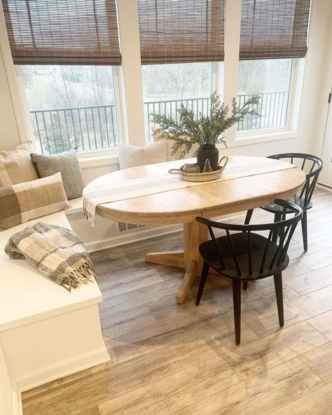 Woven shades accessorize three windows in this cozy country kitchen. The wood flooring and table set the tone for the rest of the room and provide a neutral base to display decorative pieces. If a circular pedestal table doesn't offer enough space, switch it out for an oval table that is longer and has more area. Banquette Seating In Kitchen Oval Table, Breakfast Nook With Low Windows, Banquette With Oval Table, Kitchen Nook Table Ideas, Small Kitchen With Table Ideas, Oval Dining Table With Banquette, Oval Breakfast Nook Table, Small Tables Ideas, Breakfast Nook With Windows