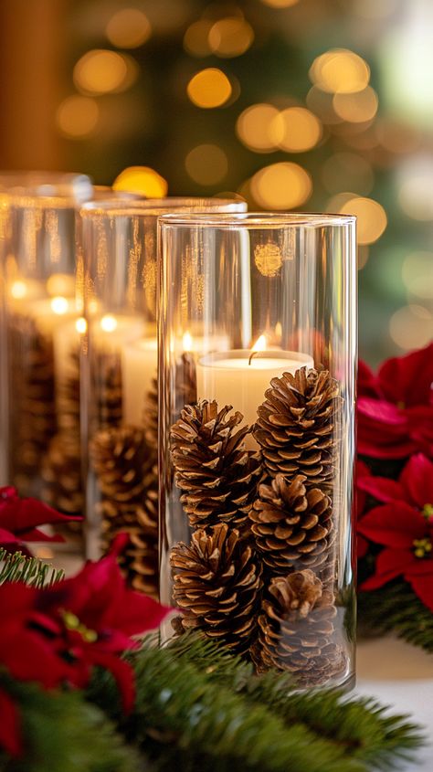 Glass candle jars with pine cones on a table adorned with red and green flowers, creating a cozy Christmas ambiance. Glass Cylinder Vases Christmas, Christmas Decor Glass Vase, Xmas Candle Decorations, Glass Bowl Christmas Centerpiece, Trendy Christmas Decor 2024, Christmas Votives Ideas, Easy Christmas Table Centerpieces Budget, Christmas Ornament Centerpieces, Christmas Vase Filler Ideas