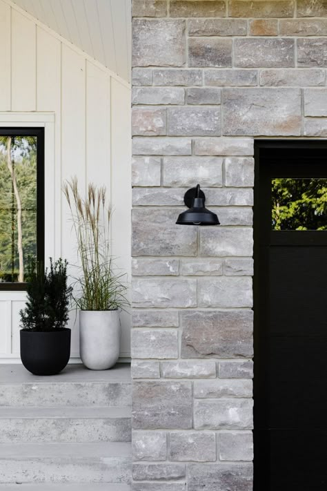 Black garage door. Farmhouse gooseneck sconce wall light. Stone and white board and batten exterior. Board And Batten Farmhouse, White Siding Exterior, White Board And Batten Siding, Black Brick Wallpaper, White Board And Batten, Farmhouse Sconces, Black Garage Door, White Farmhouse Exterior, House Colors Exterior