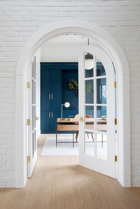 White brick frames arched French doors leading into a home office styled with a brown leather desk chair placed on a white rug at a wood and brass desk lit by a Hicks pendant. Home Office French Doors, Office French Doors, Green Home Offices, Arched French Doors, Den Library, White French Doors, Wood And Metal Desk, Home Office Shelves, Library Office