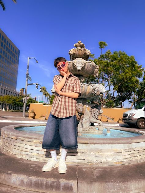 Asian boy wearing a plaid brown shirt sleeve and jnco shorts and hello kitty glasses, posing for the camera next to an elegant fountain Shorts Flannel Outfit, Jnco Jeans Outfit Men, Mens Y2k Fashion Summer, Jnco Shorts Outfit, Flannel Shorts Outfit Men, Jorts Outfit Idea Men Y2k, Men Outfits Vintage, Jorts Mens Outfits Y2k, Y2k Summer Outfit Men