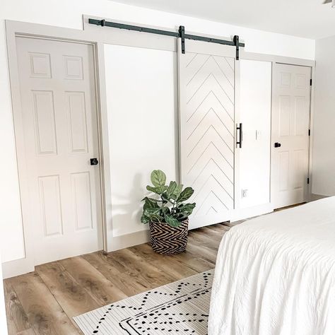 The neutral color palette of this bedroom showcases white walls and light grey trim. A light grey barn door with a chevron pattern is hung on the wall with black iron hardware. The room also features hardwood floors, a white throw rug with a modern black pattern, and a white bed. White Walls Grey Trim, Chevron Barn Door, Grey Painted Rooms, Barn Door For Bathroom, Door For Bathroom, Bedroom Barn Door, White Baseboards, Agreeable Gray Sherwin Williams, White Shiplap Wall