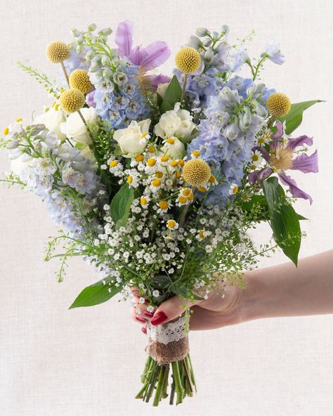 Classic garden blooms including blue delphinium, lilac clematis, daisies, and white spray roses create a charming freshly picked look, with trailing baby’s breath and greenbell adding a delicate finish. The perfect bouquet for summer weddings 🩵  Order here: https://www.appleyardflowers.com/weddings/bridal-bouquets/english-meaow-bridal-bouquet  #appleyardlondon #appleyardweddings #englishweddings #londonflorist #weddingflorist #bridetobe #weddingflowers #weddinginspiration Bluebells Wedding Bouquet, Daisy And Blue Bouquet, Light Blue Delphinium Bouquet, Blue Wildflower Wedding Theme, Blue And Lilac Wedding Flowers, Bridal Bouquet Daisies, Lavender And Daisy Bouquet, Periwinkle Flower Bouquet, Baby Blue Wedding Bouquet