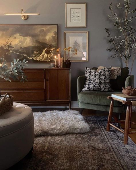 A distinguished essence is displayed within this living room, where earth tones and vintage furniture create a warm atmosphere. Gray walls welcome the company of framed images and a linear sconce. A vintage rug lays over dark wood flooring and hosts a circular ottoman and upholstered armchair...   Image: steph.homebody Room Ideas With Dark Wood Furniture, Dark Wooden Furniture Living Room, Dark Brown Wood Floors Living Rooms Gray Walls, Light Wood Furniture With Dark Floors, Dark Earth Tone Living Room, Sultry Living Room, Vintage Moody Living Room, Dark Flooring Living Room, Dark Wood Floors Living Room Furniture