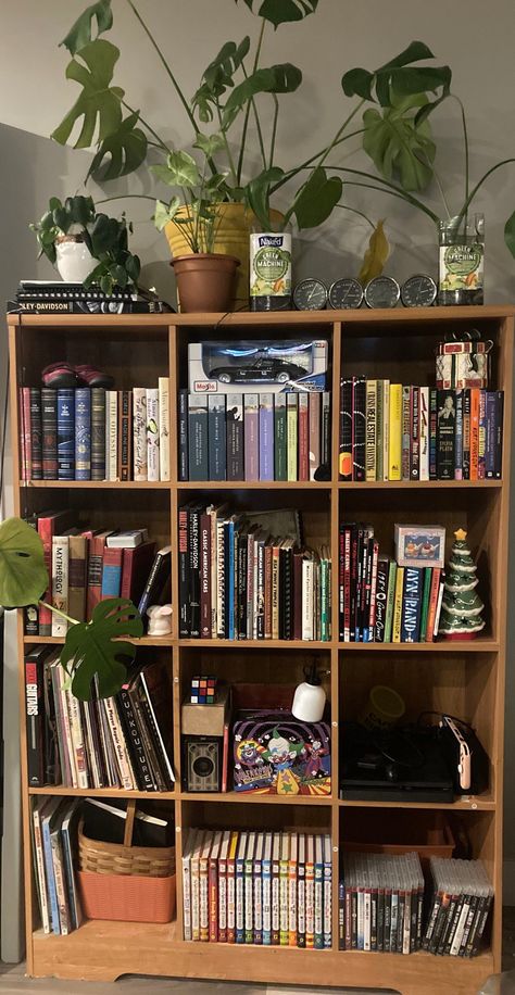 Light brown bookshelf. The bookshelf is one of those grided ones where each shelf is a square. 4 squares tall, 3 squares wide, 12 squares in total. fairly neatly organized. Lots of books and video games on their own compartments of the shelf. One shelf has a cute basket. A couple shelves have some plants, there is also larger plants ontop of the book shelf, aswell as some decorative coins? It looks very pretty overall. Mildly cottagecore. Open Shelf Bookshelf, Bookshelf Maximalist, Diy Vintage Bookshelf, Book Case Decorating Ideas Display, Brown Bookshelf Aesthetic, Wooden Bookshelf Aesthetic, Small Apartment Library, Trinket Bookshelf, 70s Bookshelf