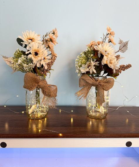 Are you looking for a unique and charming way to brighten up your space? Whether you are planning a wedding, an engagement, or any other special occasion, or you just want to add some cozy and rustic vibes to your home, these neutral earth tone wildflower centerpieces are perfect for you! Each centerpiece features a beautiful bouquet of hand-picked artificial wildflowers in a Mason jar with lights and a burlap bow. The flowers are carefully arranged to create a natural and harmonious look, and the lights add a touch of warmth and magic. The burlap bow adds a finishing touch of elegance and simplicity. These centerpieces are available in either pint size or quart size, and you can choose from sets of 2, 4, 6, or 8. They are easy to display and move around, and they will make any room feel m Mason Jar Fairy Lights Centerpieces, Mason Jar Table Centerpieces, Rustic Centerpieces For Party, Rustic Baby Shower Centerpieces, Centerpieces Without Flowers, Rustic Wedding Centerpieces Diy, Mason Jar Centerpieces For Weddings, Growler Centerpiece Wedding, Mason Jar Fall Flower Arrangements