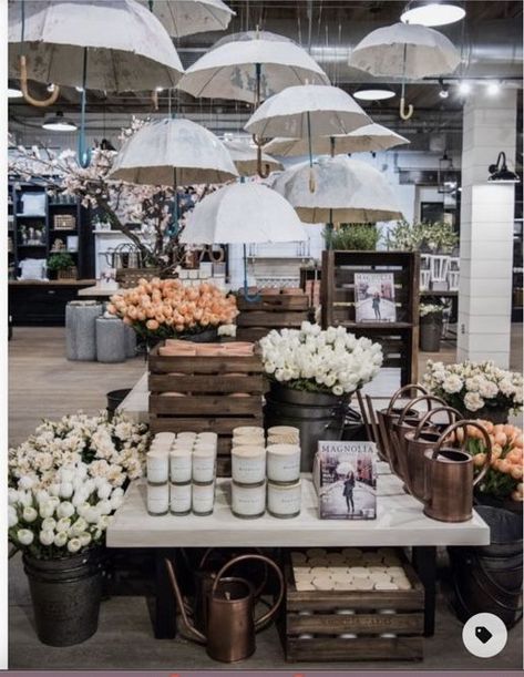 With the stunning color palette of white and orange, as well as the umbrella above the focus display, it's easy to imagine a stunning DIY fall retail display idea for this store. As a focal point, this table draws your attention to the area. The plants, candles, magazine, and water container make for a colorful and eye-catching display. This is probably the first thing I'd want to look at when I walk into the store. Boutique Window Displays, Spring Window Display, Magnolia Homes Paint, Joanna Gaines Paint, Gift Shop Displays, Window Display Retail, Focus Point, Store Window Display, Store Window Displays