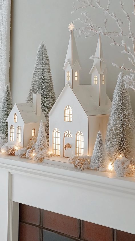 Scandinavian-style white wooden Christmas village with lighted windows, church, and trees on a fireplace shelf. White And Gold Christmas Village, White House Christmas Village, All White Christmas Village, Modern Christmas Village Display, White Ceramic Houses Christmas, White Christmas Village Display, Scandinavian Christmas Decor Ideas, White Ceramic Houses, Wooden Christmas Village