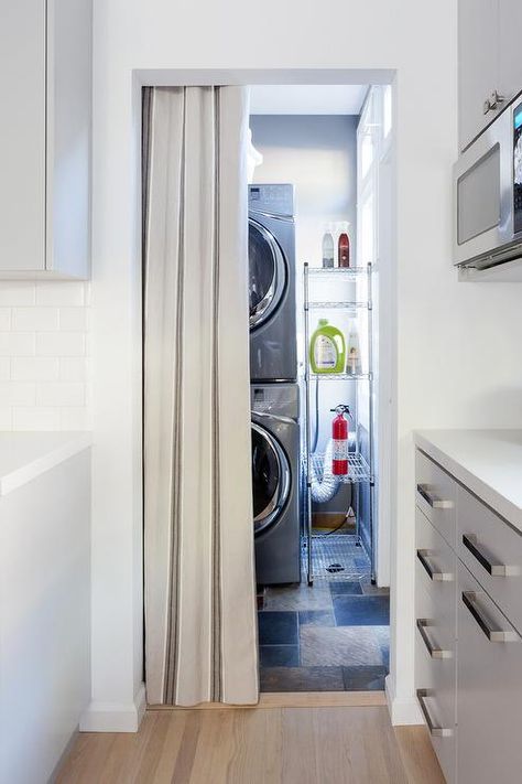 Separated from the kitchen by a gray curtain hung from a doorway, a modern laundry room boasts a stacked silver front loading washer and dryer positioned beside a stainless steel industrial shelving unit. Doors To Hide Washer And Dryer, Curtain To Hide Washer Dryer, How To Hide Laundry Machines, How To Hide Washer And Dryer In Hallway, Washer And Dryer In Closet Small Spaces, Hiding Washer And Dryer In Kitchen, Hide Washer And Dryer In Kitchen, How To Hide Washer And Dryer In Kitchen, Hiding Washer And Dryer