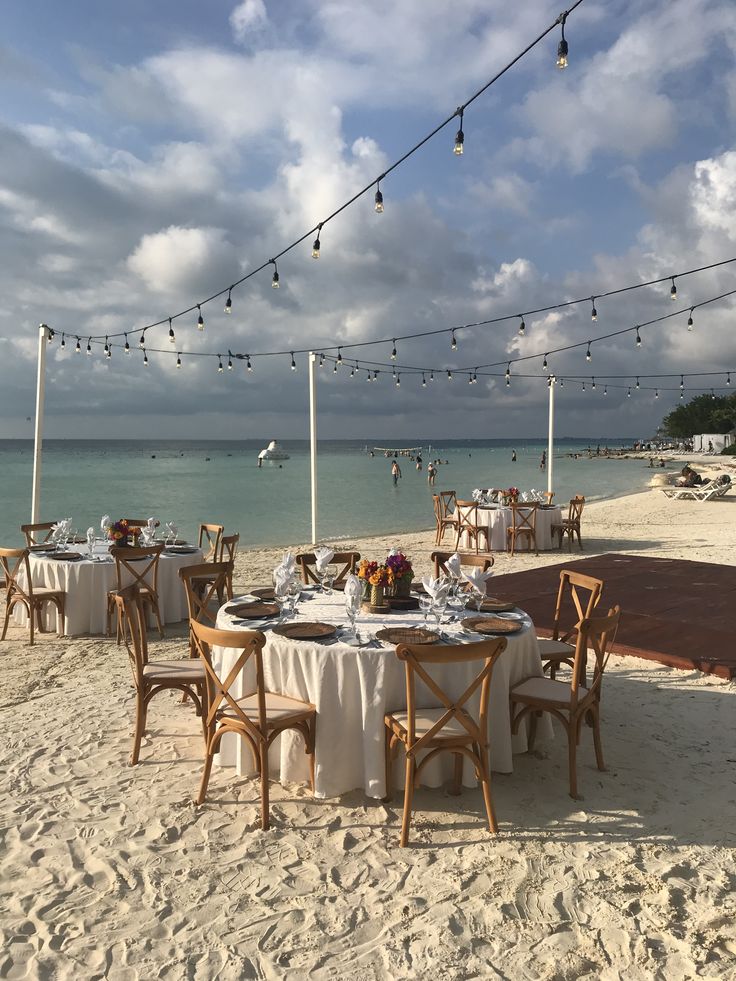 a table set up on the beach for an outdoor dining event with string lights strung over it