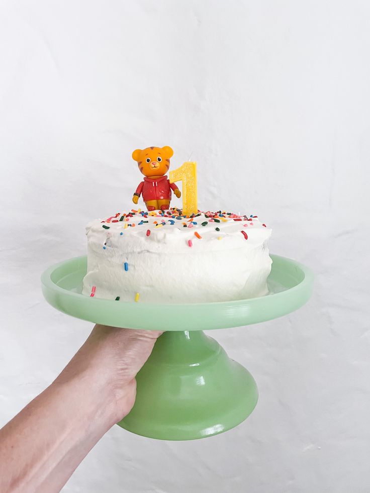 a hand holding a cake with a teddy bear on top and sprinkles