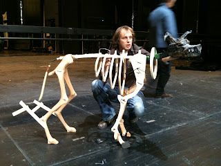 a man kneeling down next to a wooden skeleton on top of a black stage floor