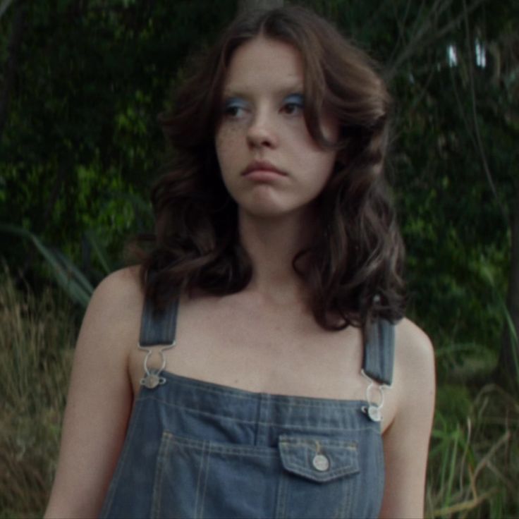 a woman with blue eyes and overalls standing in the woods