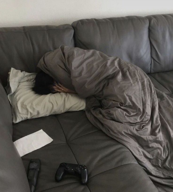 a person sleeping on top of a couch covered by a blanket and pillows with video game remotes