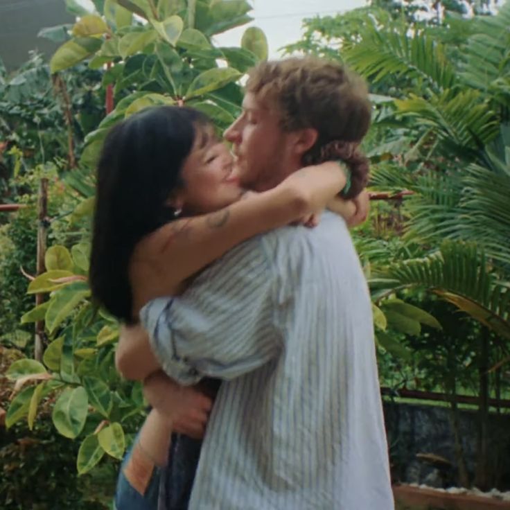 a man and woman hug each other in front of some trees with green plants behind them