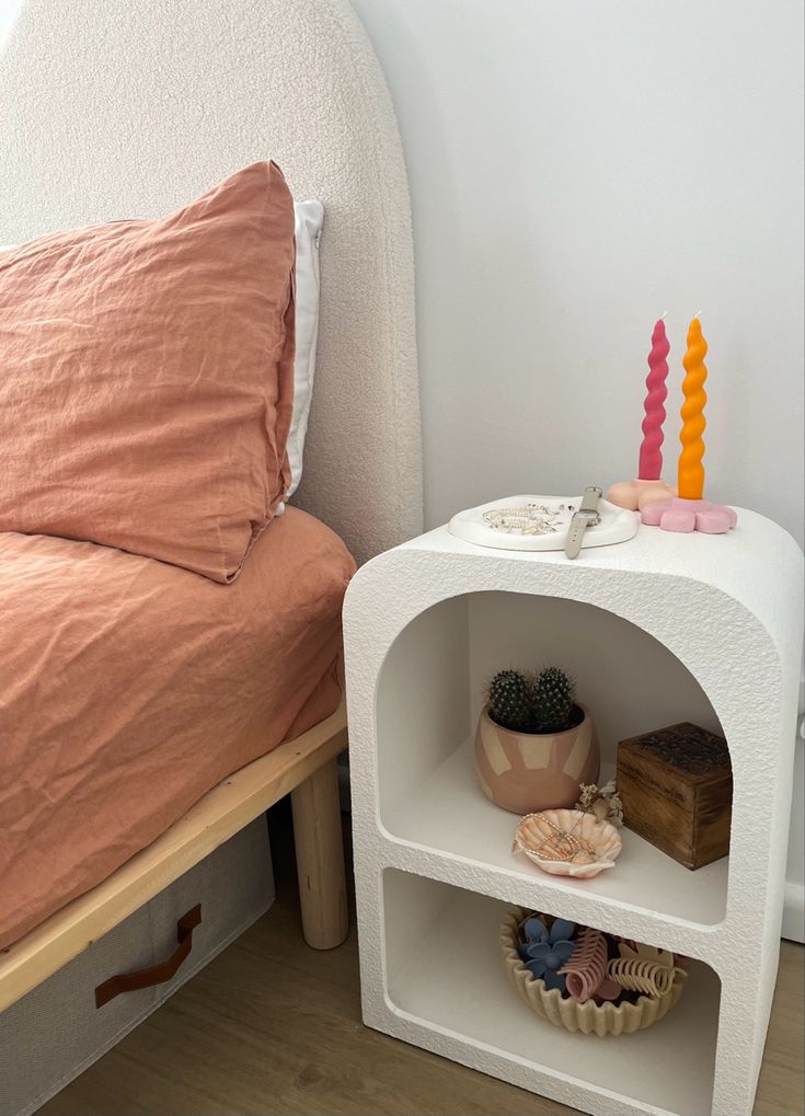 a small white shelf next to a bed with pillows and blankets on top of it