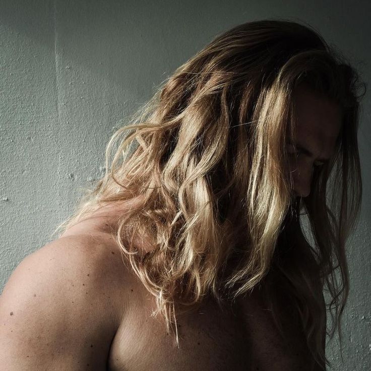 a man with long hair and no shirt standing in front of a white wall looking off to the side