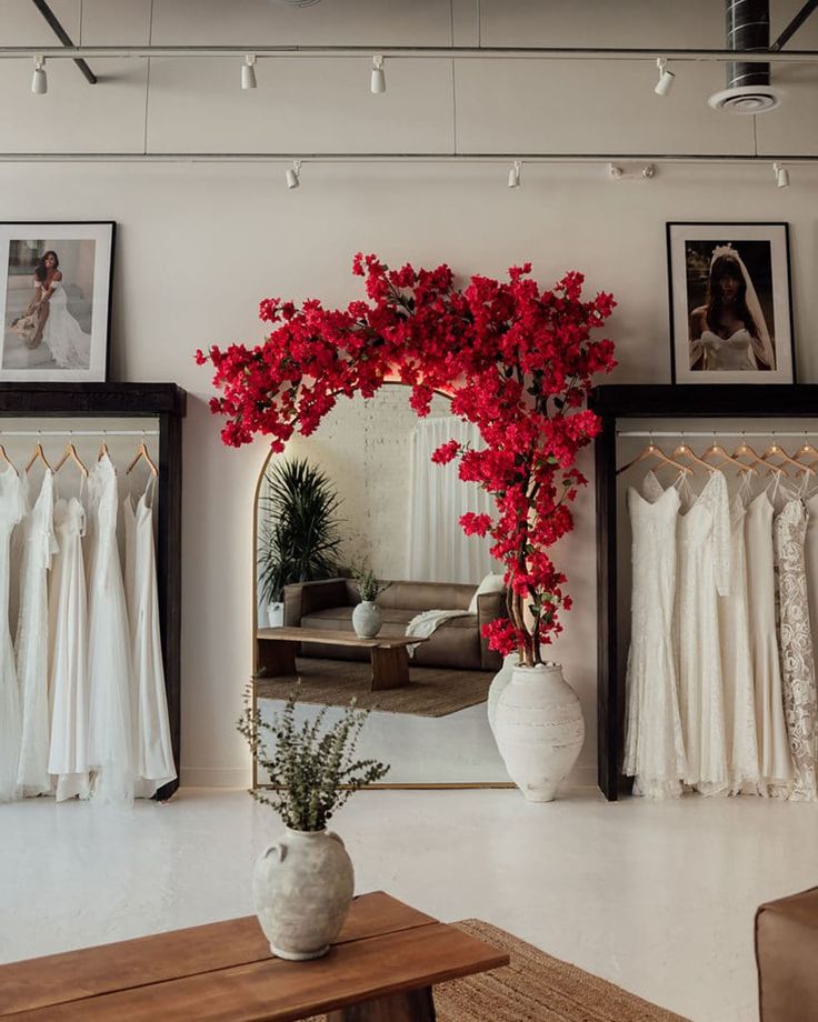 a room filled with lots of white dresses and red flowers in vases next to a mirror