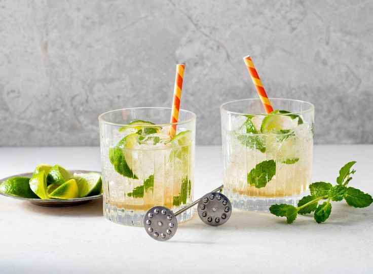 two glasses filled with ice and limes on top of a white table next to a plate