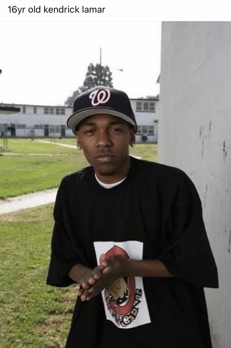 a man standing in front of a white wall wearing a baseball cap and black t - shirt