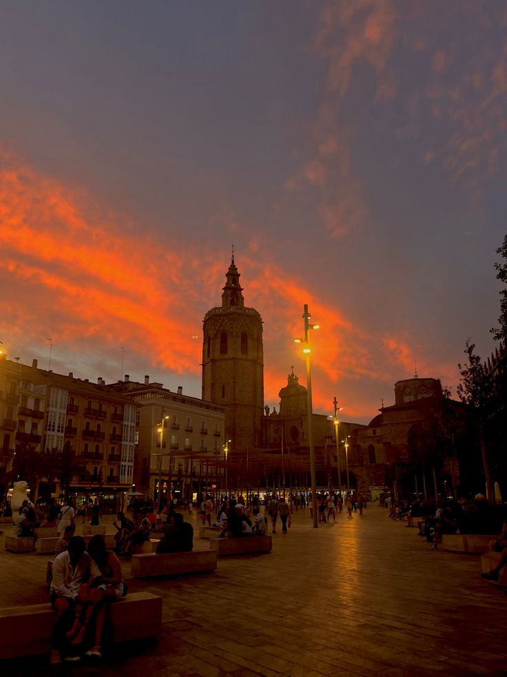 people are sitting on benches in front of a clock tower at dusk with the sun going down