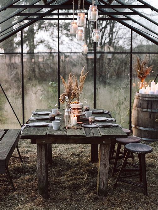 an outdoor table with candles and plates in front of a glass house filled with plants