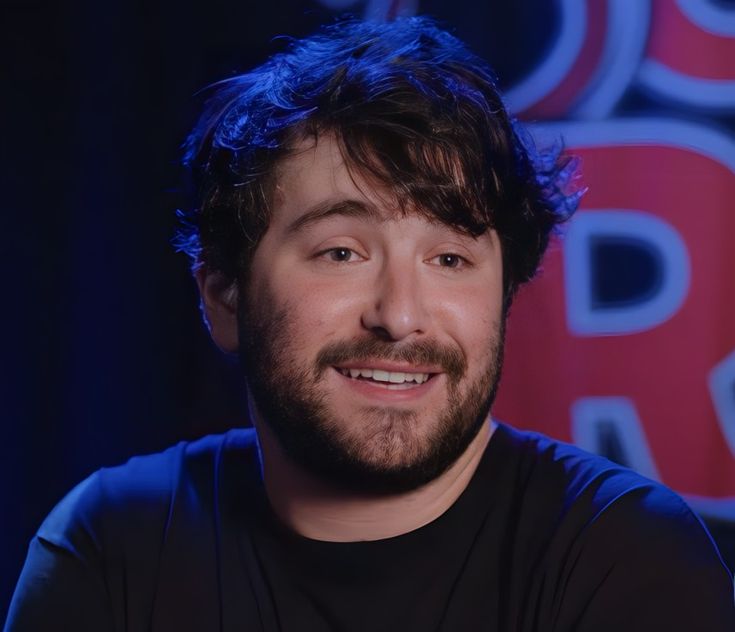 a bearded man in a black shirt is smiling at the camera with blue lighting behind him