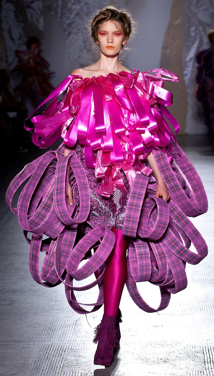 a woman in a purple dress with large pink ribbons on her shoulders and arms, walking down the runway