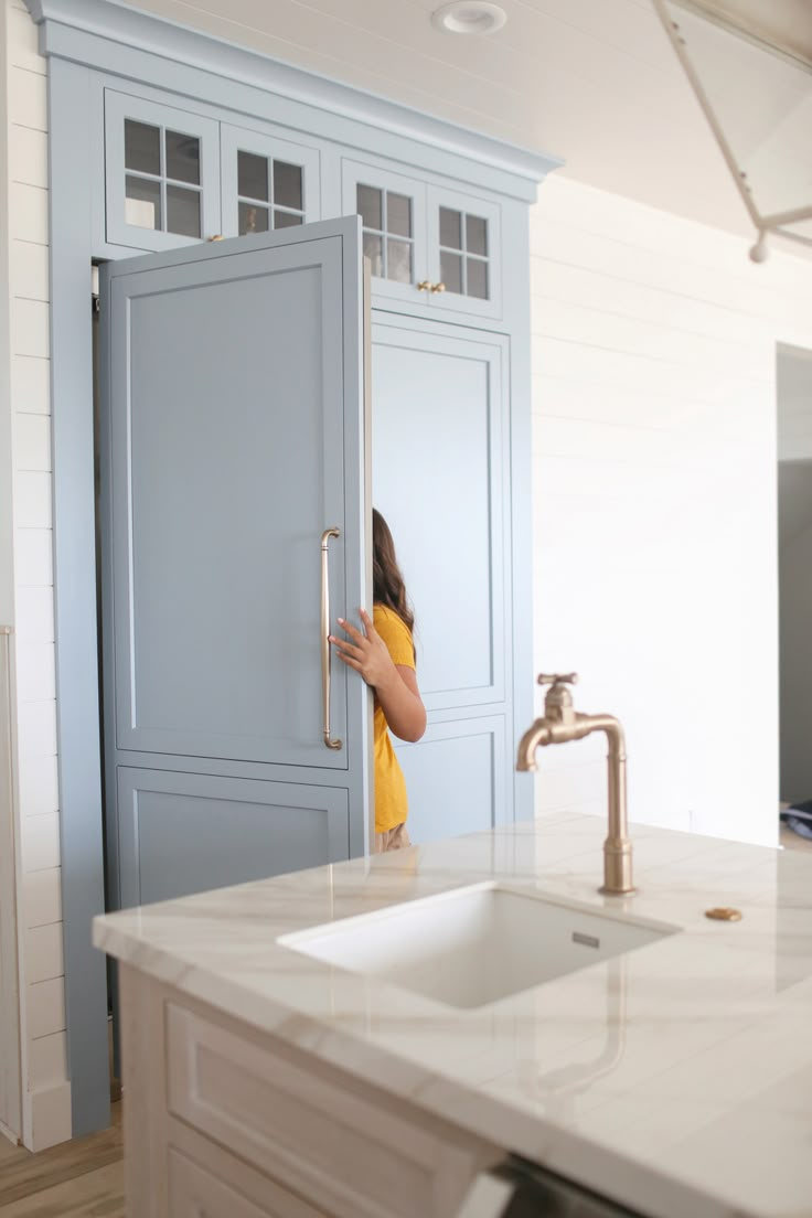 a woman peeking into a blue cabinet in a kitchen with a sink and faucet