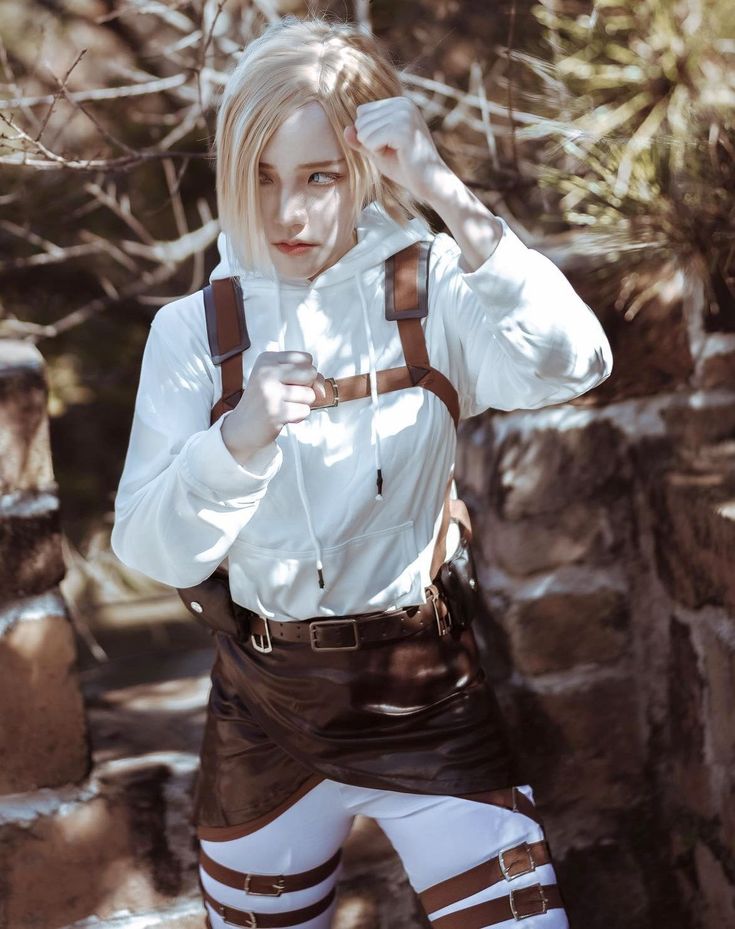 a woman in white shirt and brown suspenders standing next to stone wall with trees