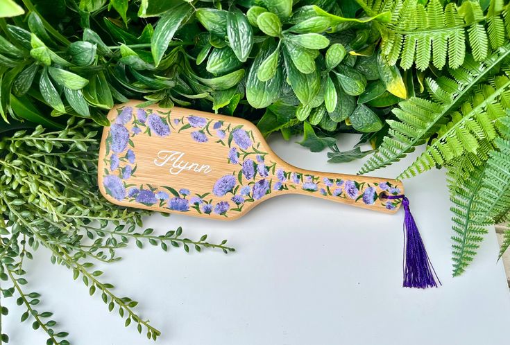 personalized wooden paddle with purple flowers and tassels on it next to greenery