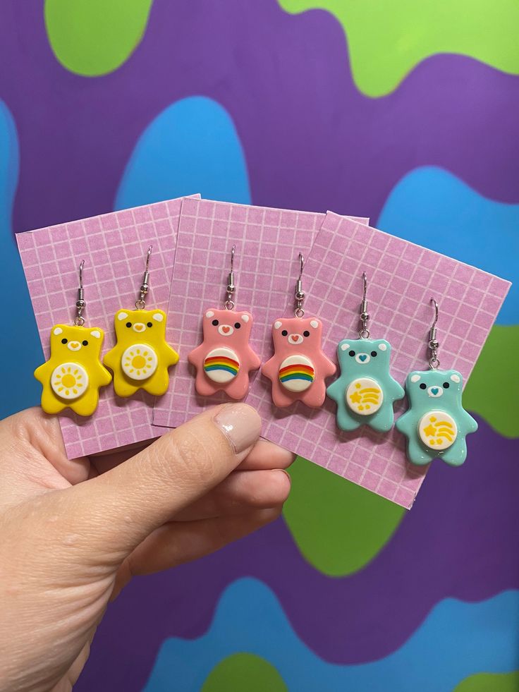 a person holding five small bear earrings in front of a purple background with blue, green, yellow and pink colors